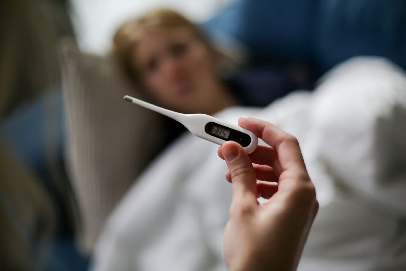 Hand holding a digital thermometer near a sick person resting in bed